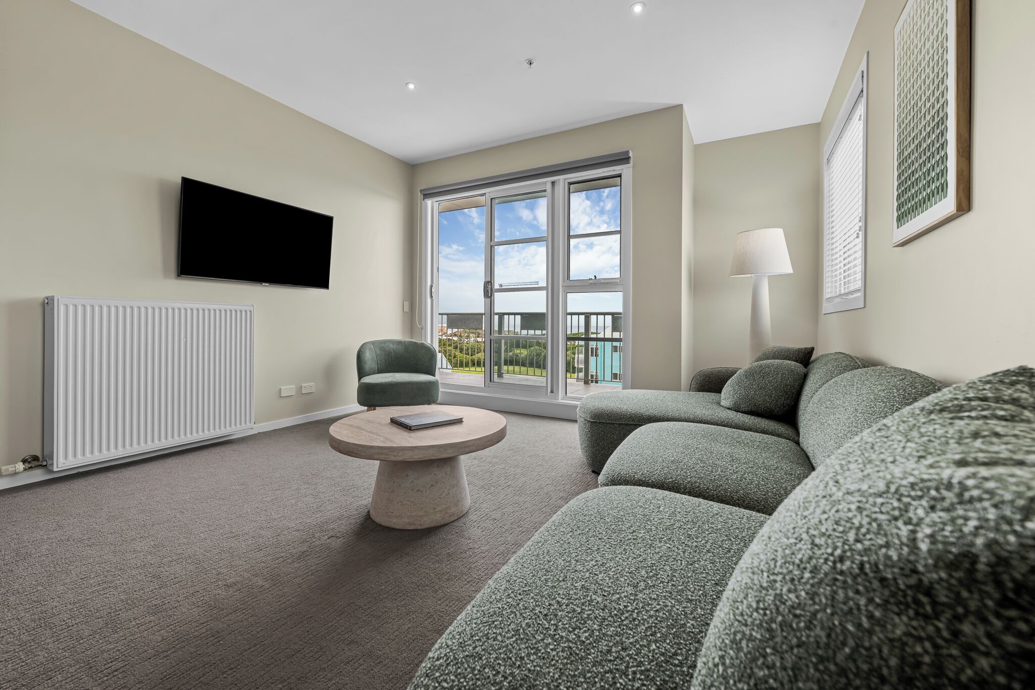 Een moderne woonkamer met een grijze bank, een ronde salontafel, een wandgemonteerde televisie en een groot raam met uitzicht op groen.