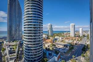 Un edificio alto con fachada curva, rodeado de otros edificios altos, un estacionamiento y vista al mar.