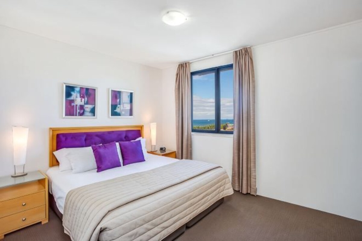 A hotel room with a bed, bedside tables, a window with curtains, and two wall-mounted pictures.