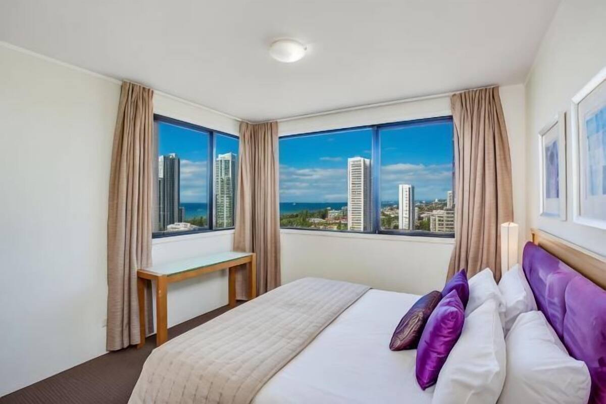 A hotel room with a bed, a window with curtains, a view of buildings, and a cityscape.