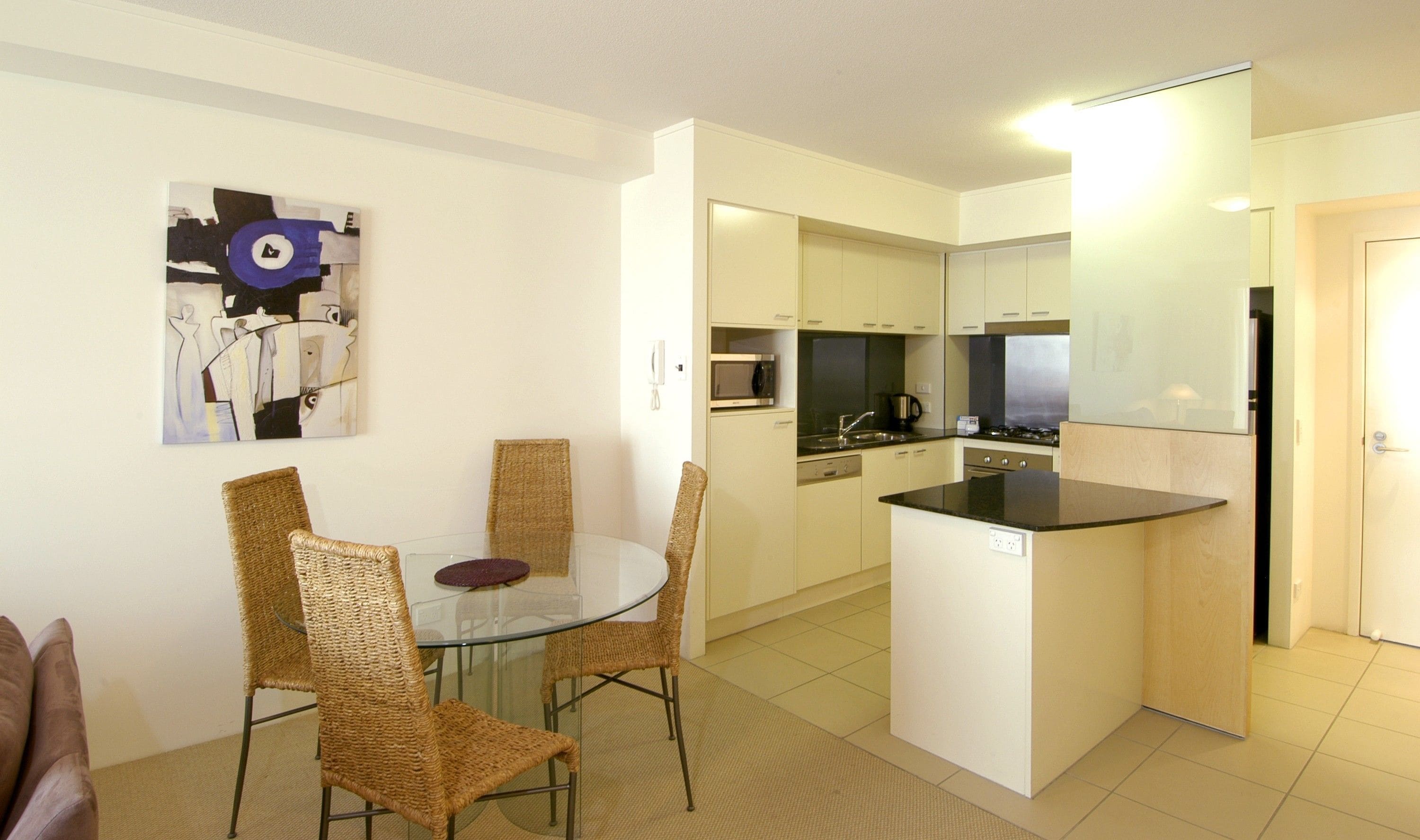 A compact kitchen with a microwave, stove, and sink. There is a dining area with wicker chairs and a glass table. A painting is on the wall.