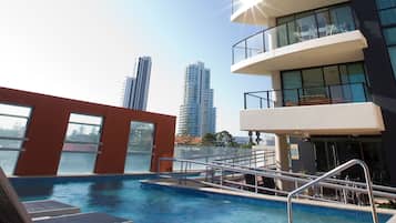 Una piscina en la azotea con vistas despejadas a edificios altos, barandilla de cristal y un balcón soleado.
