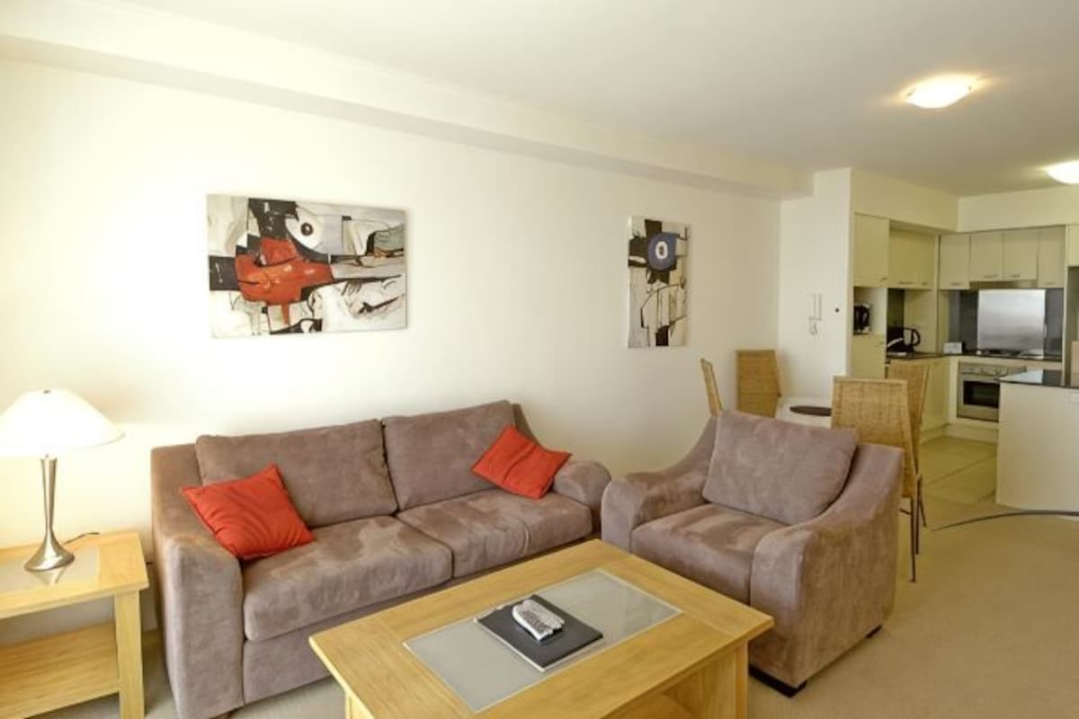 A living room with two sofas, a coffee table, and a lamp. There is a kitchen in the background with a TV and a refrigerator.