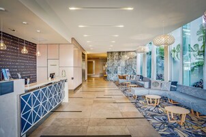 A modern hotel lobby with a reception desk, seating area, and large windows with plant views.