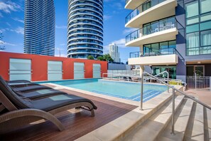 Un área de piscina en la azotea con sillones reclinables, piscina y edificios altos al fondo.