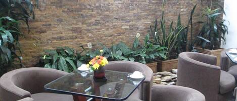 Un área de descanso con pared de ladrillo, varias plantas en macetas y una mesa de vidrio rodeada de sillones marrones.