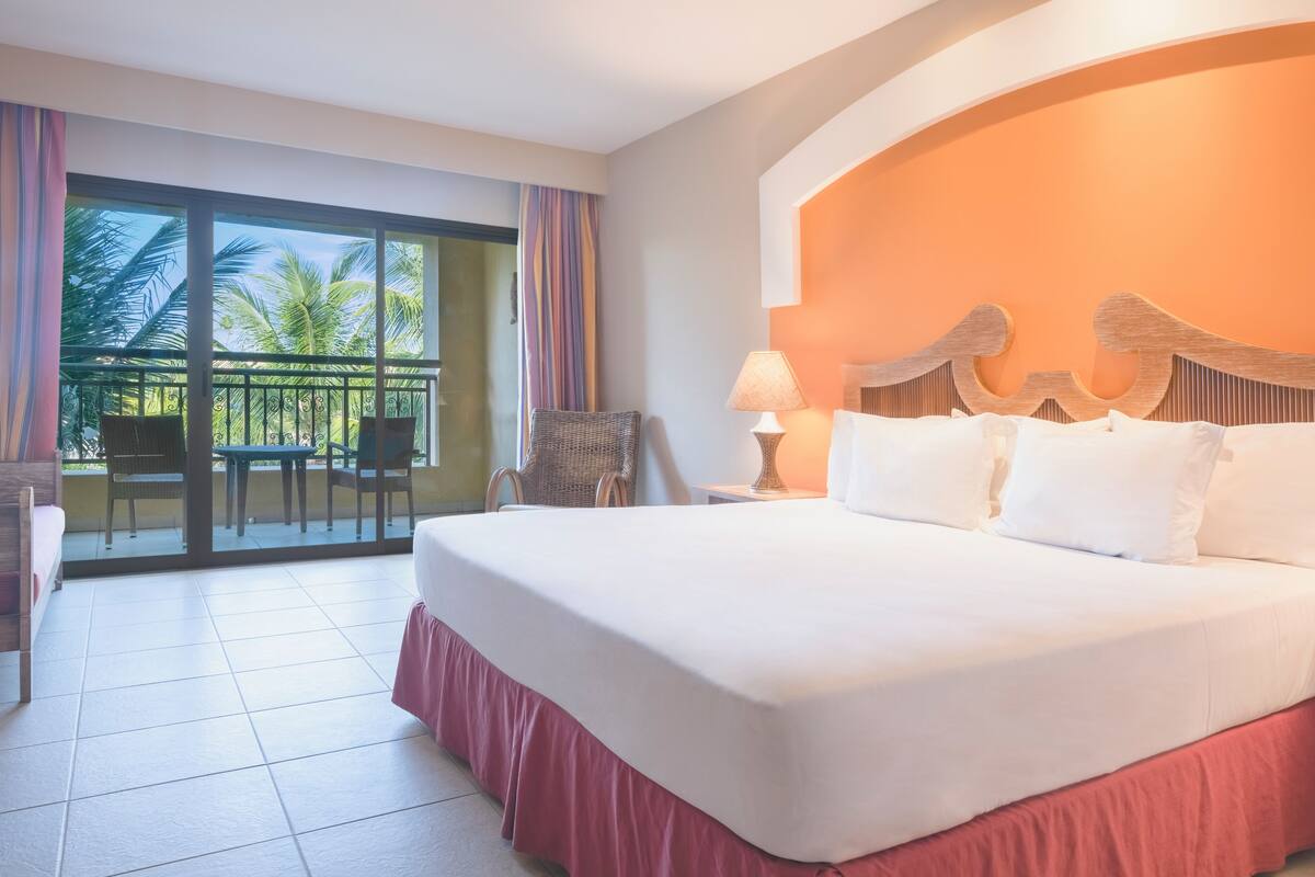Une chambre d’hôtel avec un grand lit, un balcon avec des chaises et une vue sur les palmiers.
