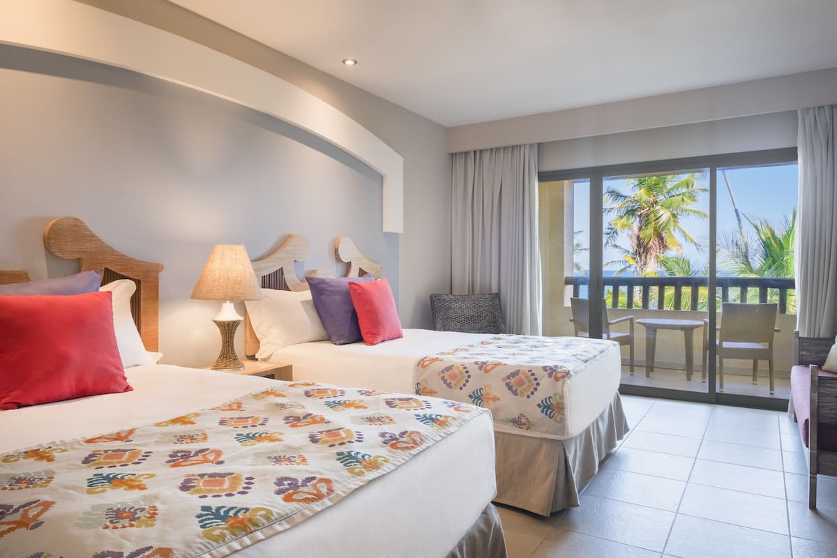 Une chambre d’hôtel avec deux lits, un balcon donnant sur des palmiers, ainsi qu’une table et une chaise sur le balcon.