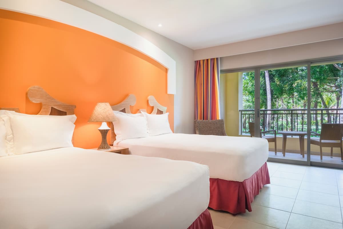 Une chambre d’hôtel avec deux lits, un balcon donnant sur la verdure et une grande fenêtre avec des rideaux.