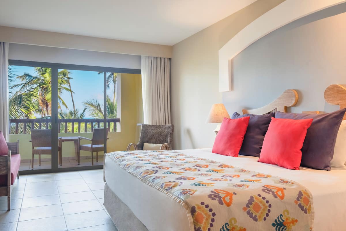 Une chambre d’hôtel avec un grand lit, un balcon avec des chaises et une vue sur les palmiers.