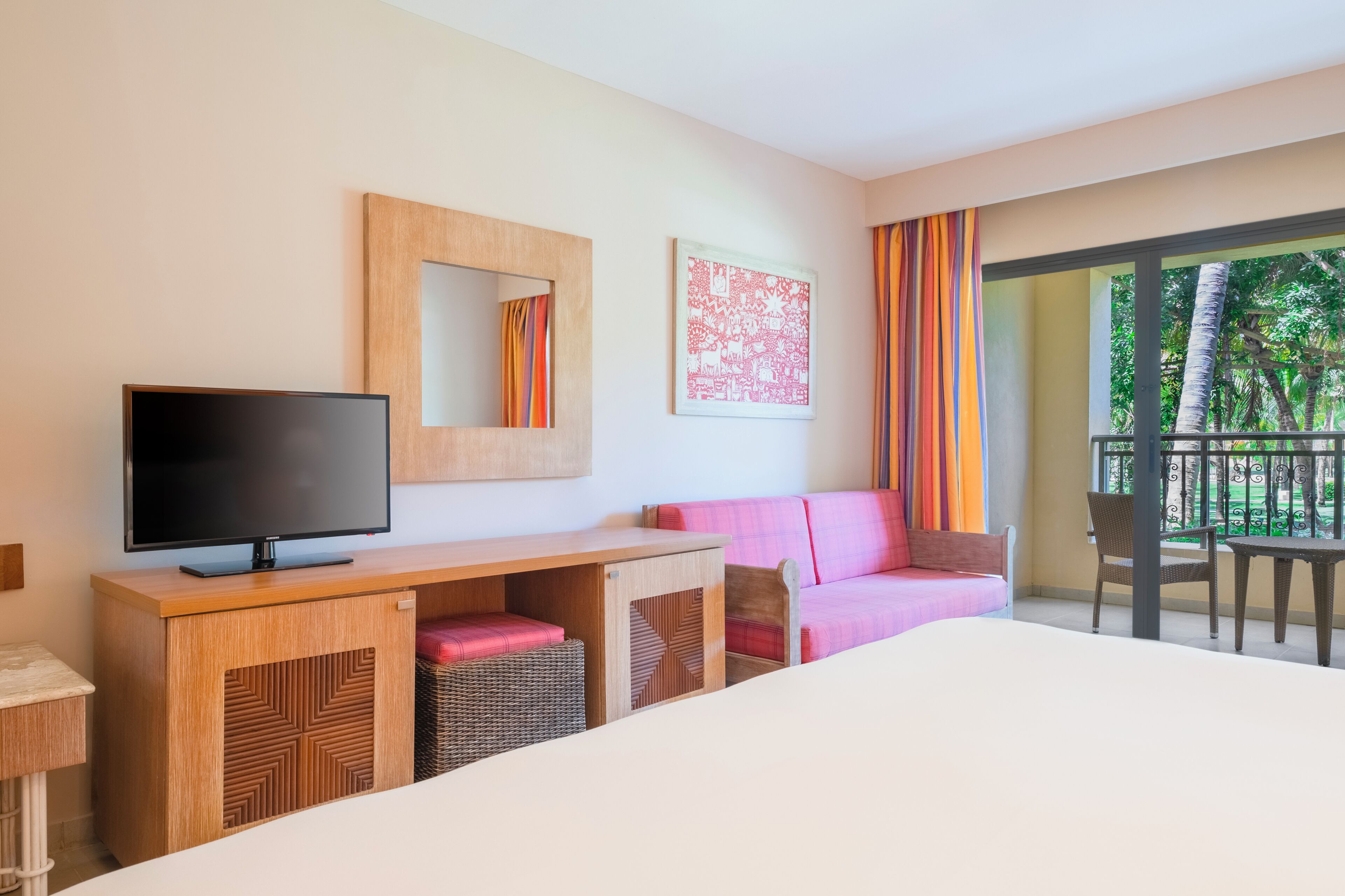 Une chambre d’hôtel équipée d’une télévision, d’un bureau en bois, d’un canapé rose et d’un balcon offrant une vue sur la verdure.