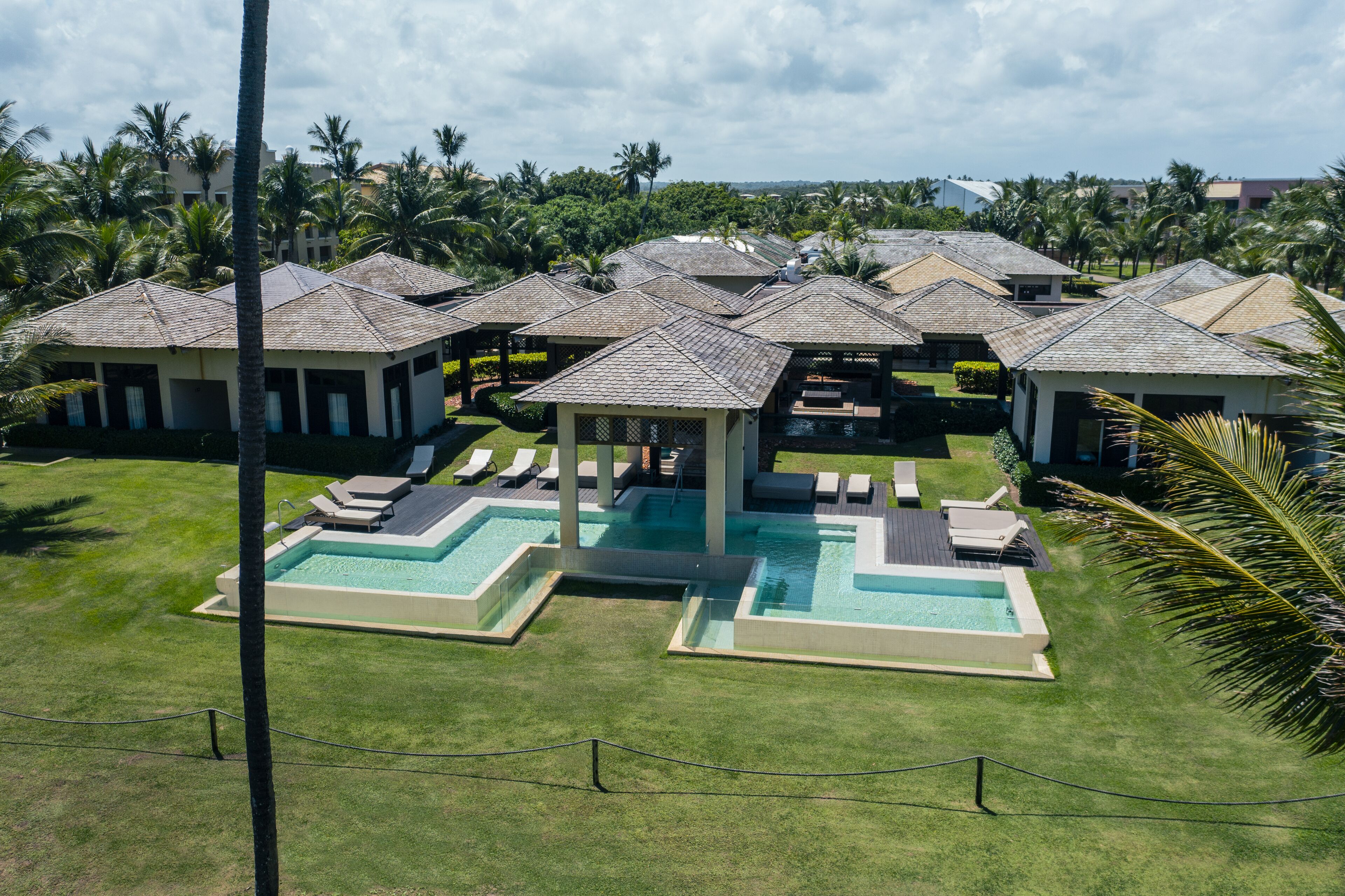 A resort with multiple bungalow-style buildings, a central pool area with lounge chairs, and surrounding palm trees.