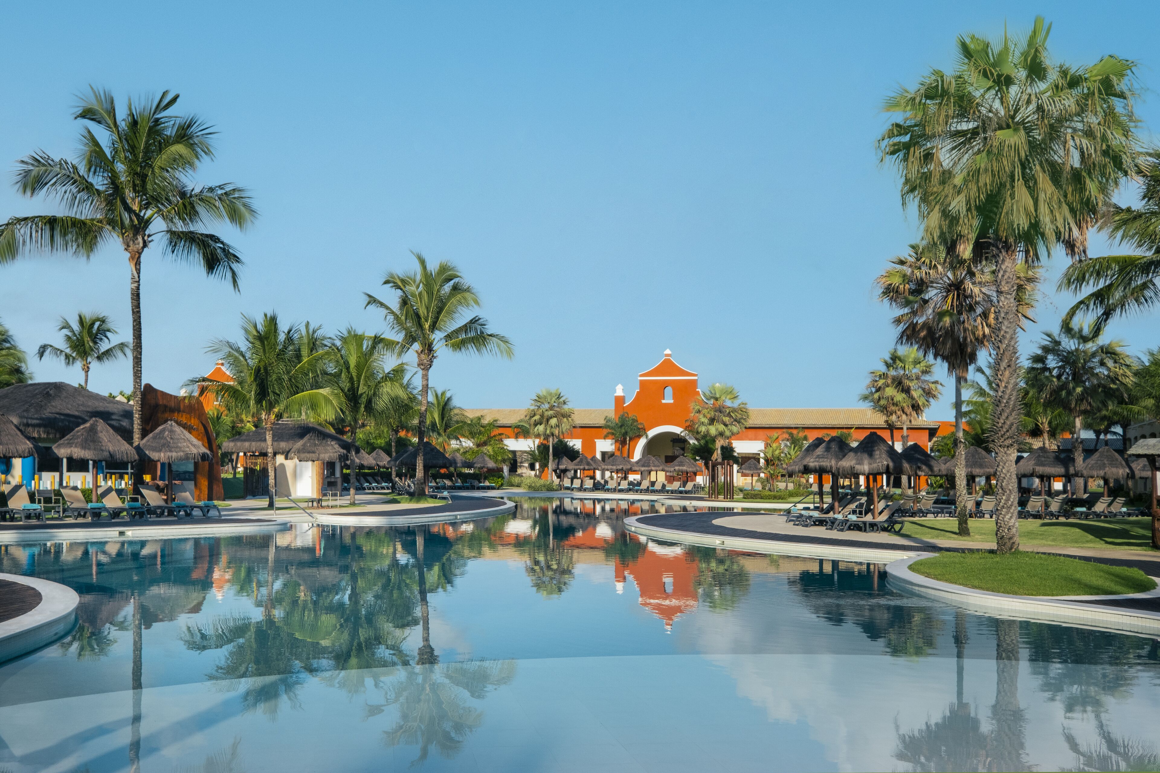 Um resort com uma piscina grande, palmeiras e um edifício de fachada laranja.