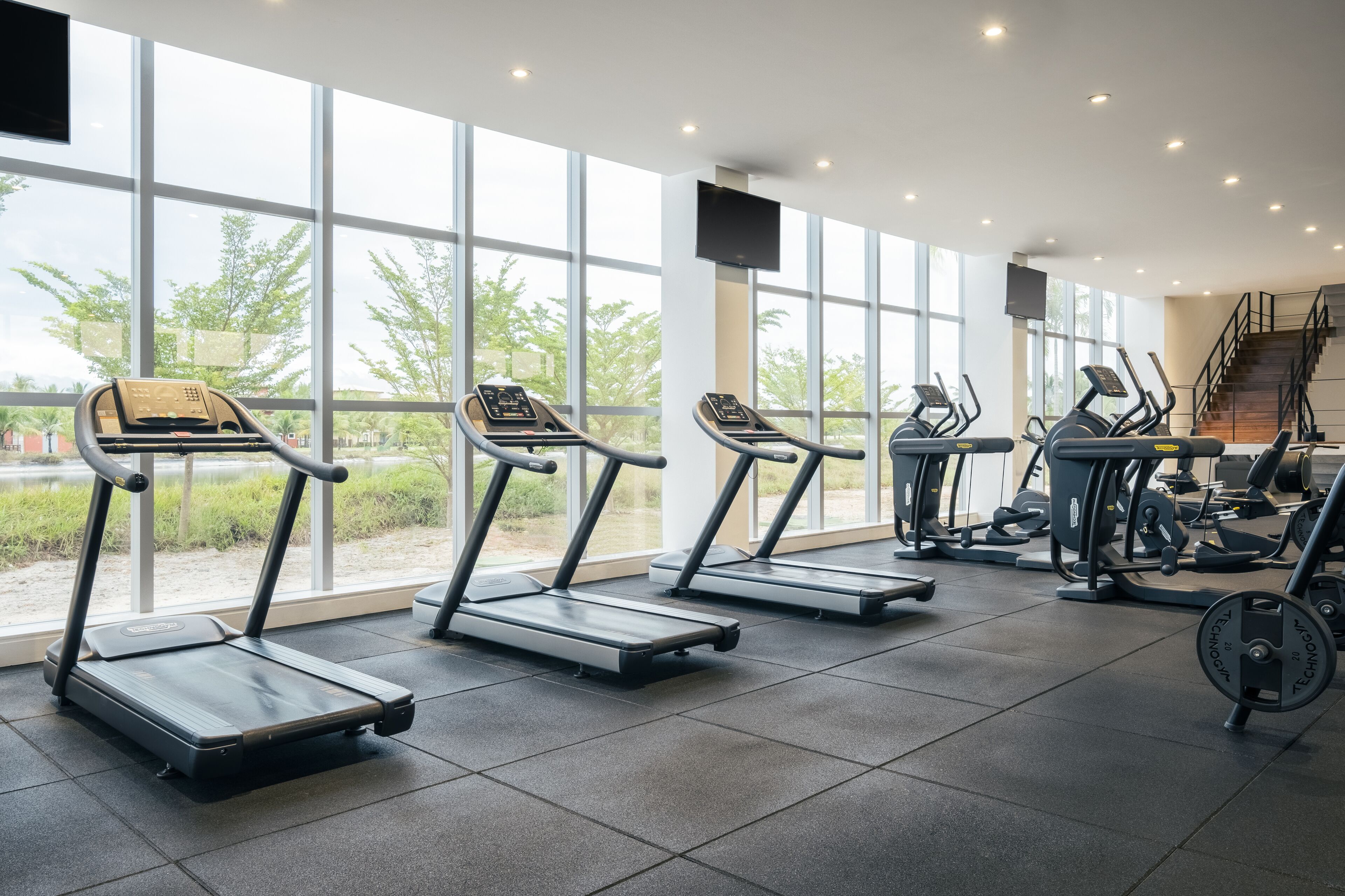 Une salle de sport équipée de tapis de course, d’appareils de musculation et dotée d’une grande fenêtre donnant sur un paysage arboré.
