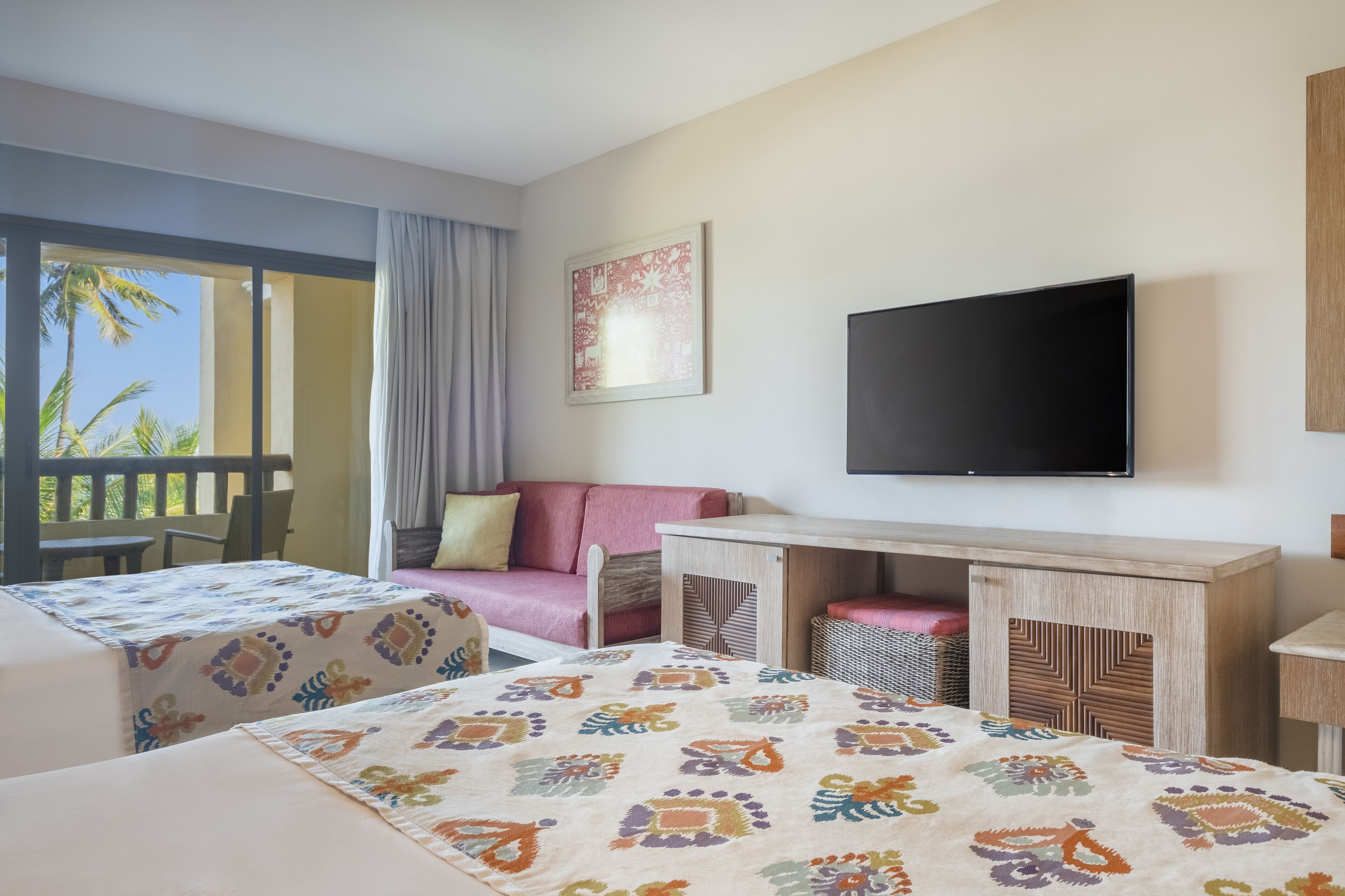 Een hotelkamer met een bed, een bureau, een televisie en een balkon met uitzicht op palmbomen.
