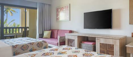 A hotel room with a bed, a desk, a television, and a balcony with a view of palm trees.