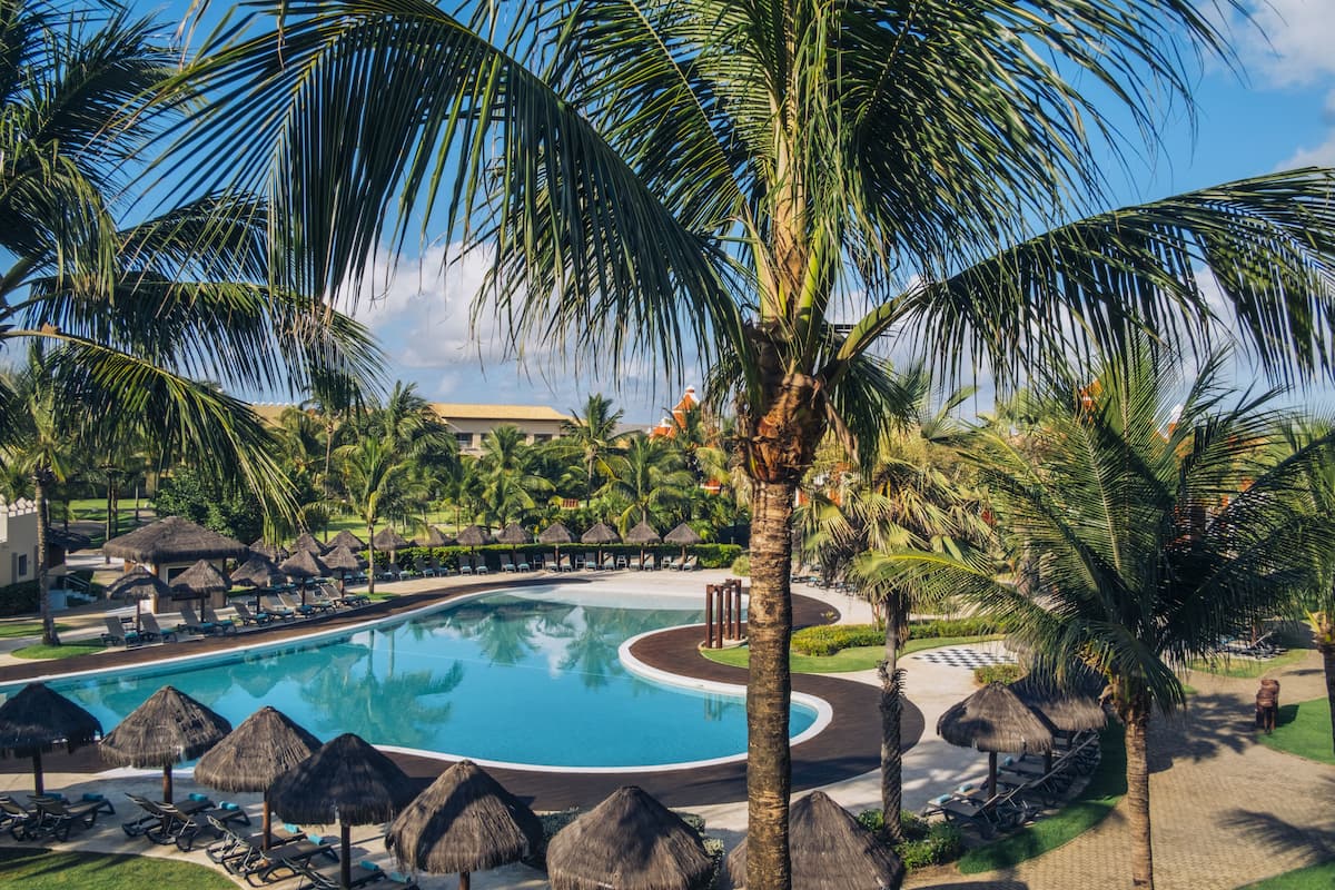 Une piscine d’un complexe hôtelier, entourée de palmiers et ombragée par des parasols en feuilles de palmier, avec des chaises longues et un ciel d’un bleu limpide.