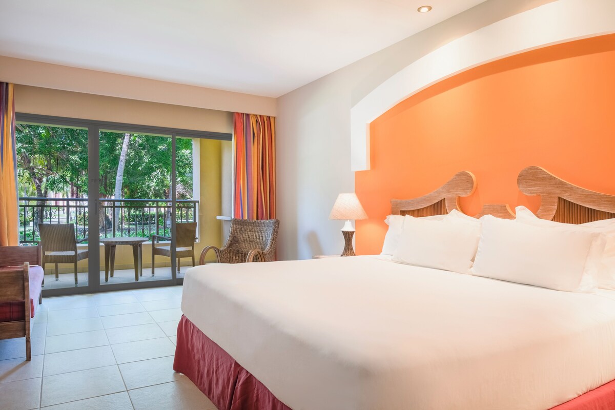 A hotel room with a large bed, a balcony with wicker chairs, and a view of greenery.