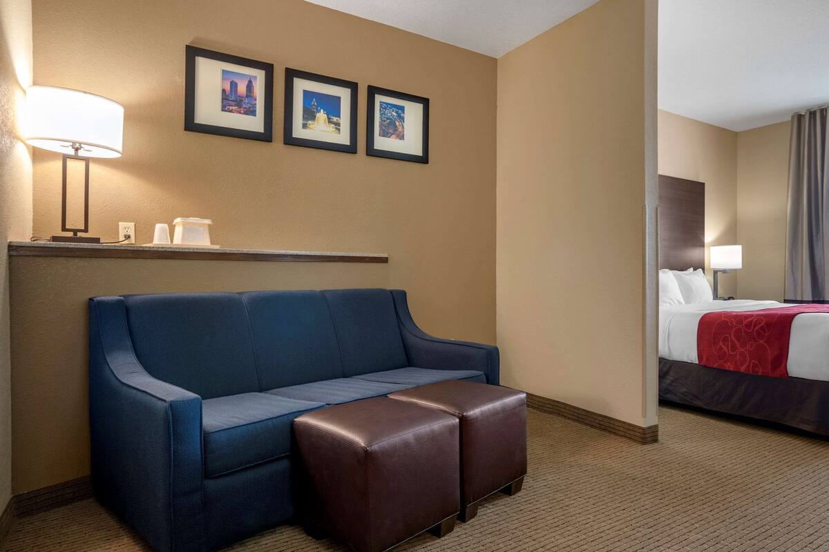 A hotel room with a blue sofa, a brown ottoman, a bed with a red blanket, a nightstand with a lamp, and three framed pictures on the wall.