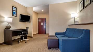A hotel room with a desk, chair, TV, couch, and ottoman.