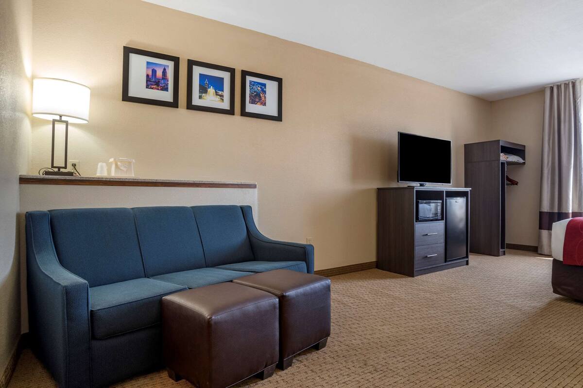 A hotel room with a blue sofa, a brown ottoman, a flat-screen TV, a minibar, and a coat rack.