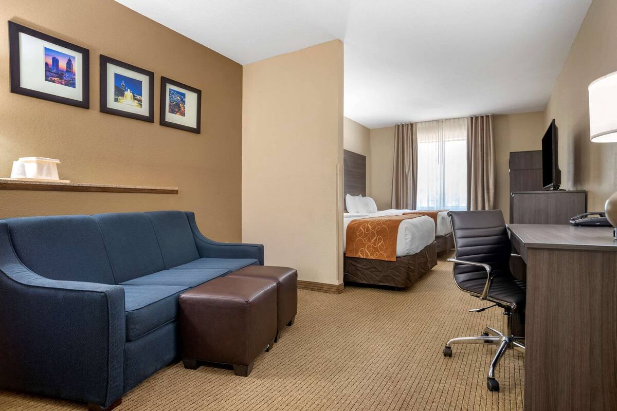 A hotel room with a blue sofa, a small brown ottoman, a desk with a chair, a bed with a white and orange bedspread, and three framed pictures on the wall.