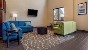 A spacious living area with a blue sofa, a green sofa, and a round ottoman. A flat-screen TV is mounted on the wall, and there is a window providing natural light. - Comfort Suites Mobile (Mobile)