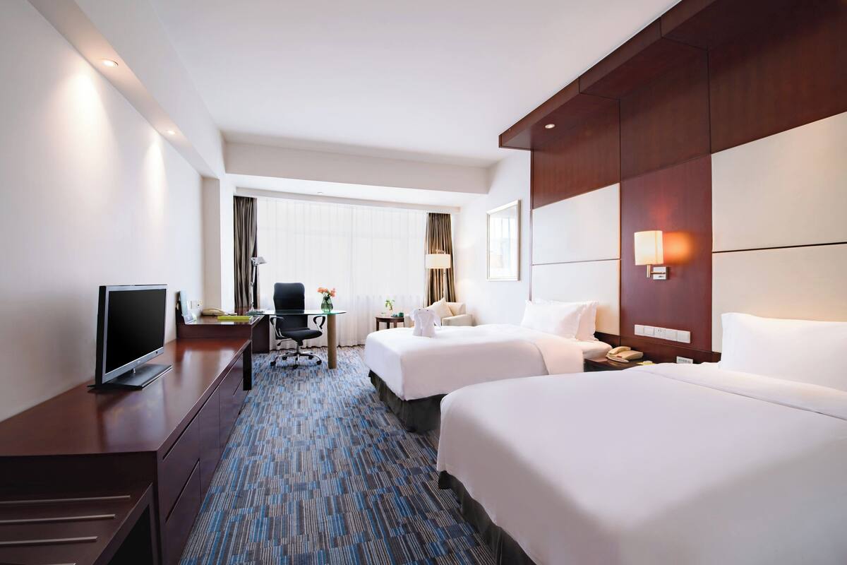 A hotel room with two beds, a desk with a television, a chair, a lamp, and a window with curtains.