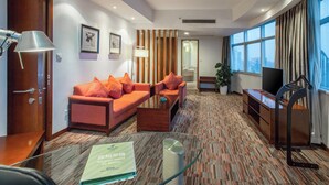 A hotel room with a sofa, armchair, coffee table, TV, and a desk with a lamp.