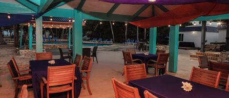 Area pranzo all'aperto con tavoli e sedie, piscina sullo sfondo e zona salotto coperta con luci a stringa.