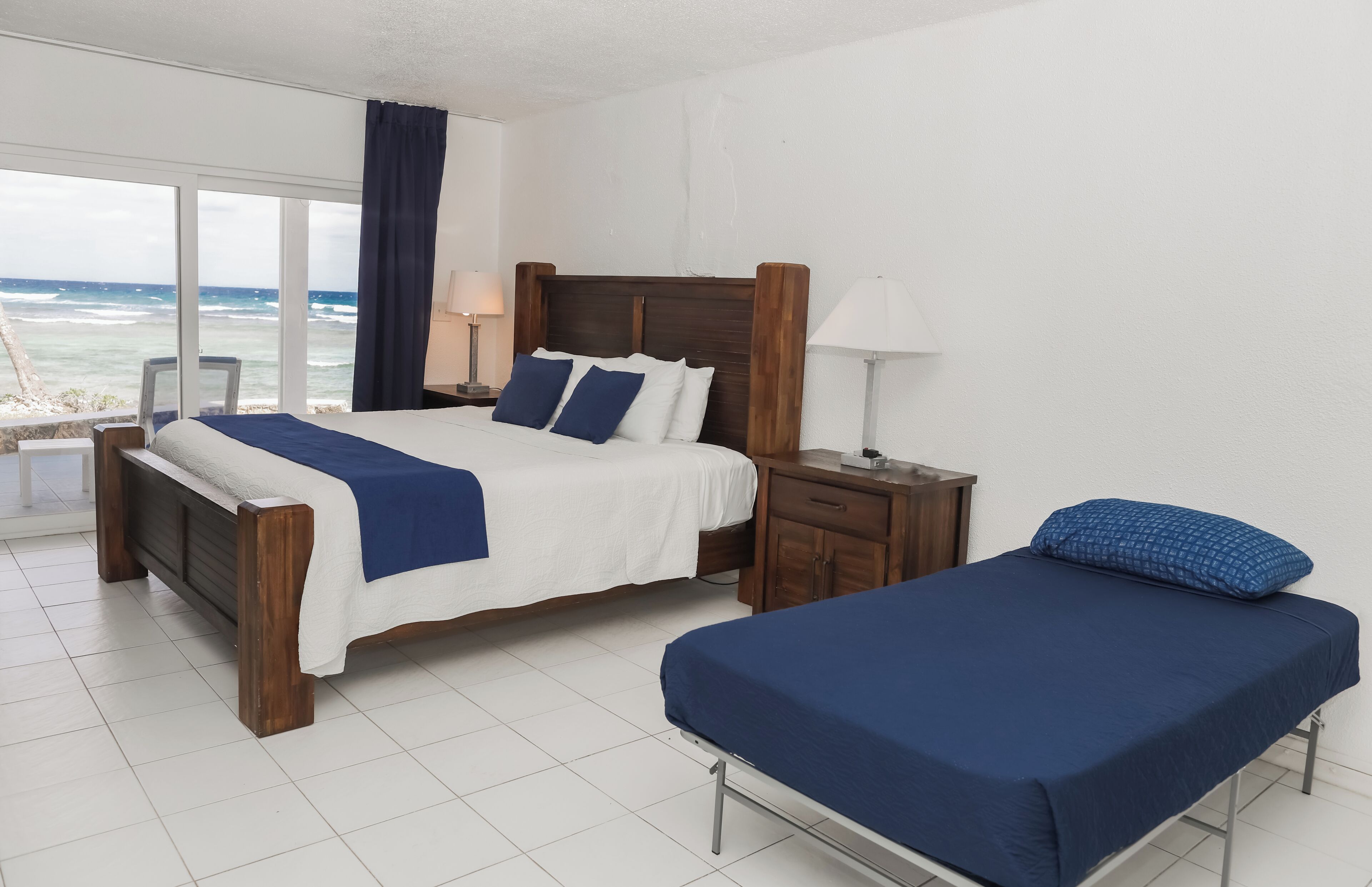 A hotel room with a large bed, a smaller bed, a nightstand, a lamp, and a view of the ocean.