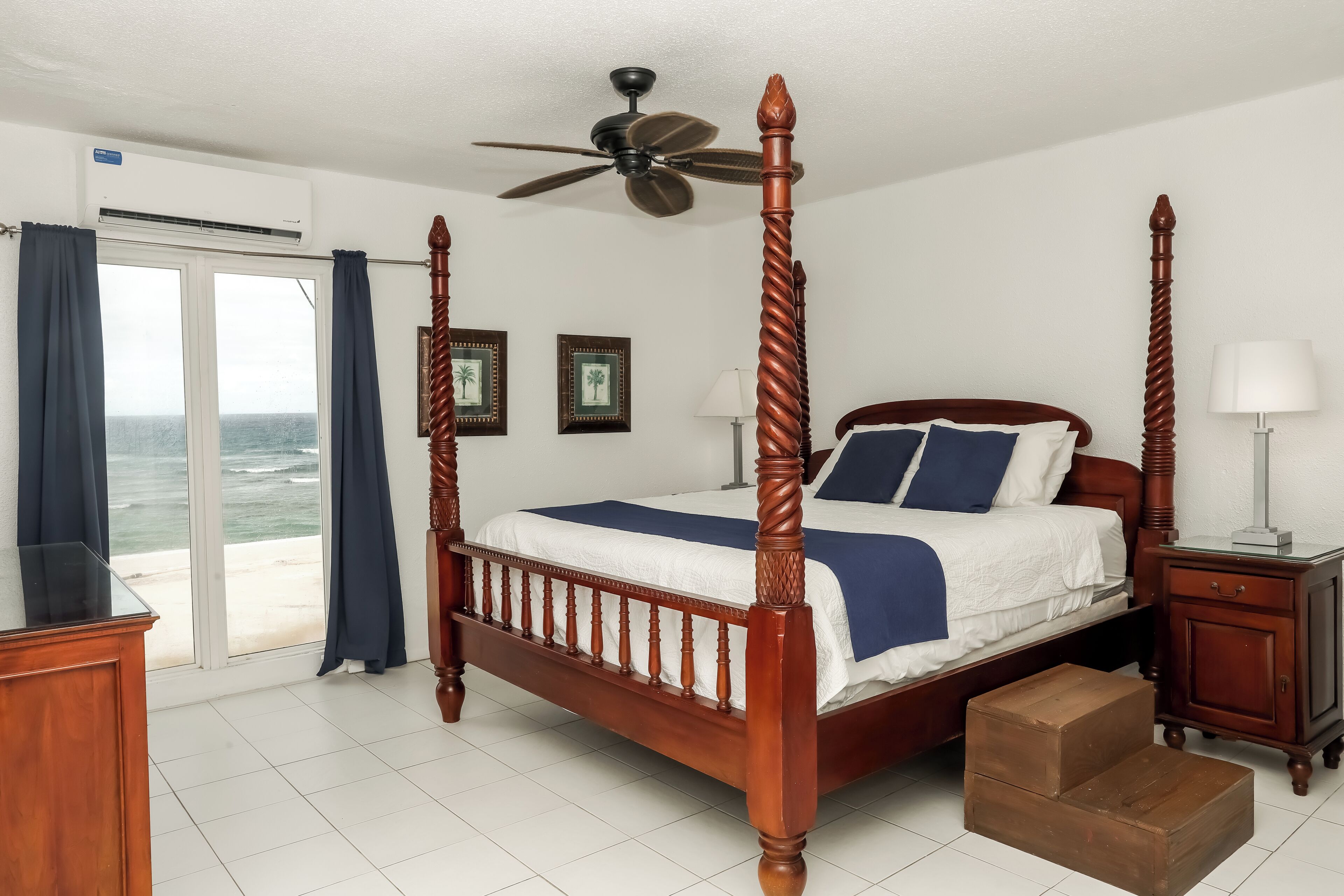 A four-poster bed with a ceiling fan, a nightstand, a lamp, and a view of the ocean through a sliding glass door.