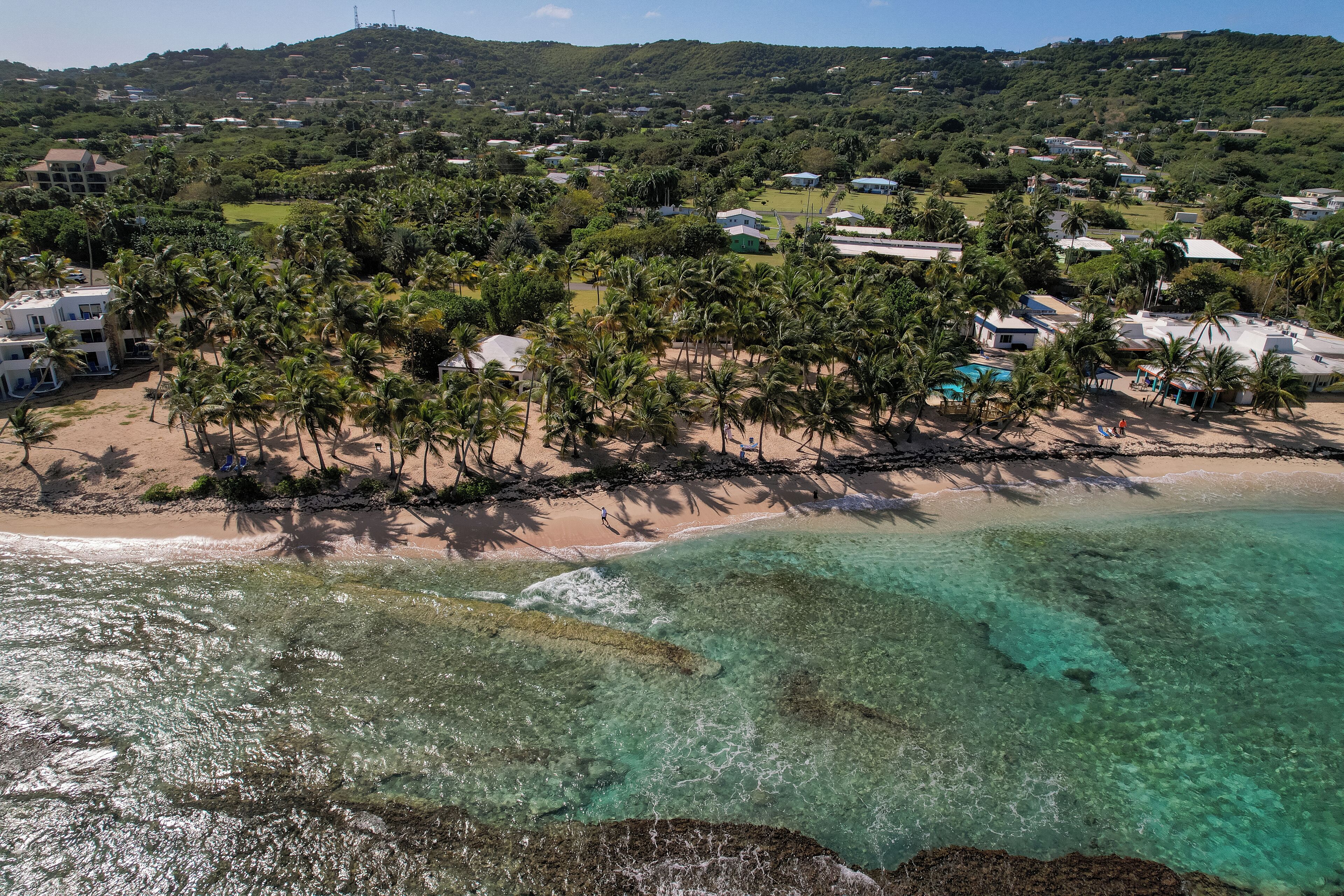 A coastal area with a beach, clear turquoise waters, and a backdrop of hills and greenery.