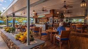 Un restaurante al aire libre con mesas y sillas de madera, una zona de bar y una jardinera de piedra con plantas tropicales.