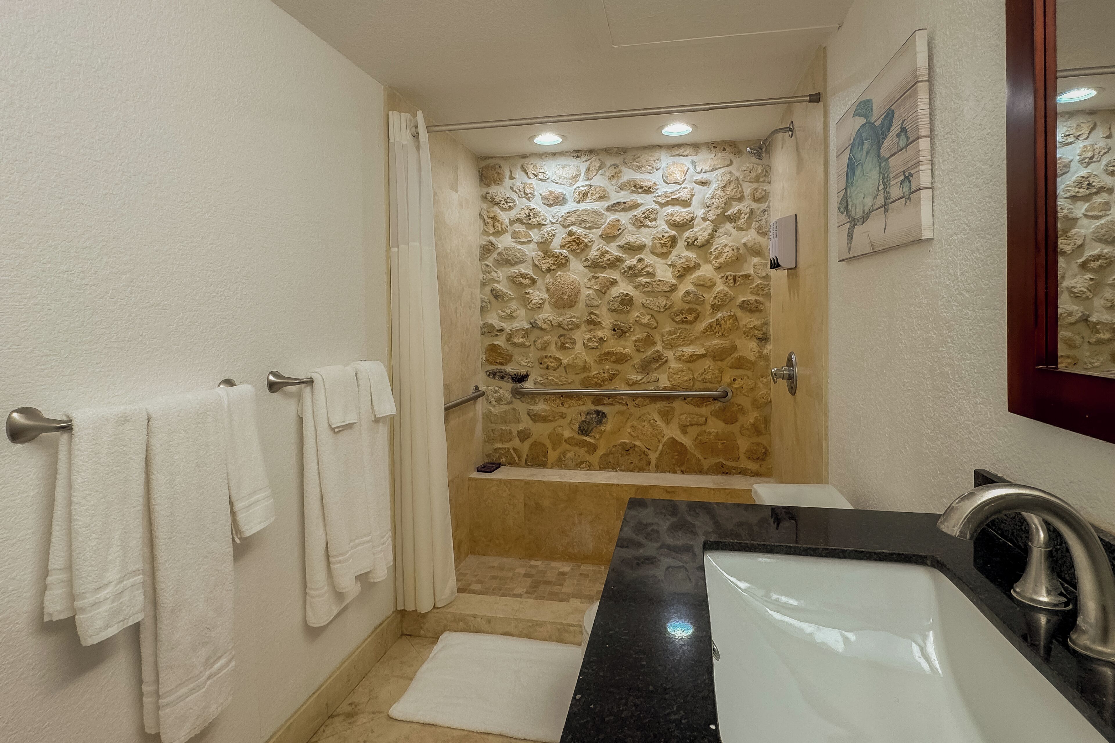 A bathroom with a stone shower wall, a bathtub, a sink with a black countertop, and white towels on a rack.