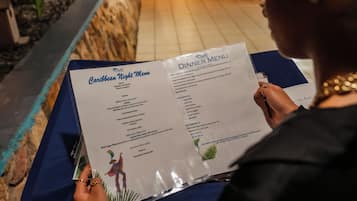 Iemand leest een menu voor Caraïbische avond in een restaurant met een blauwe tafelkleed en een accentmuur van steen.