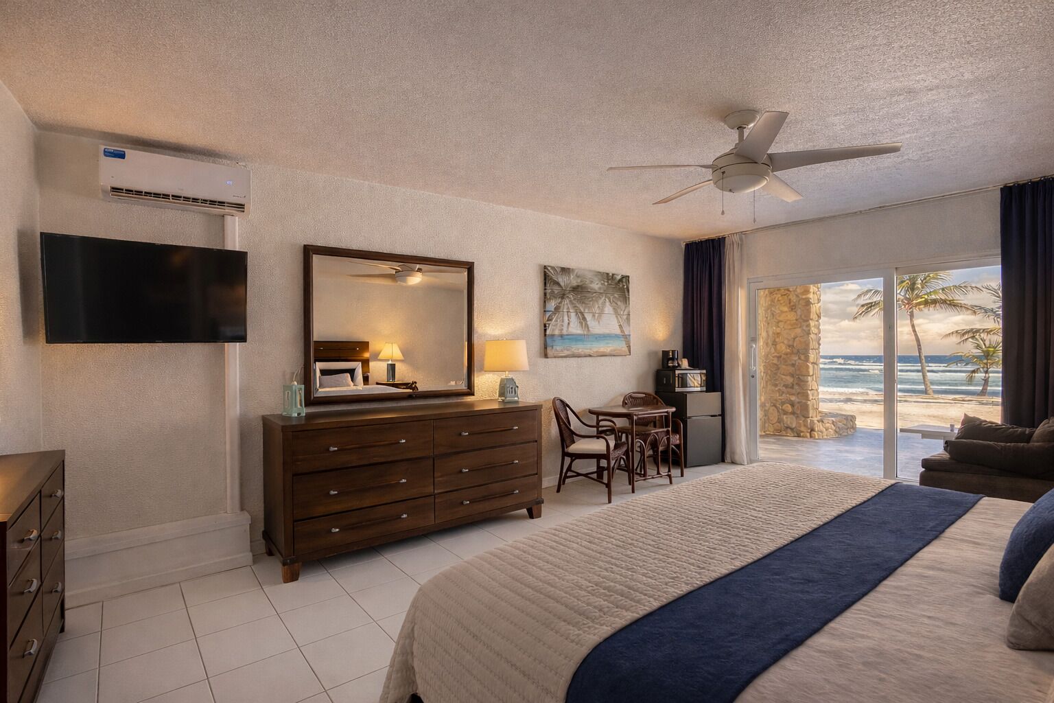 A hotel room with a bed, a desk, a chair, a television, a ceiling fan, and a view of the beach.