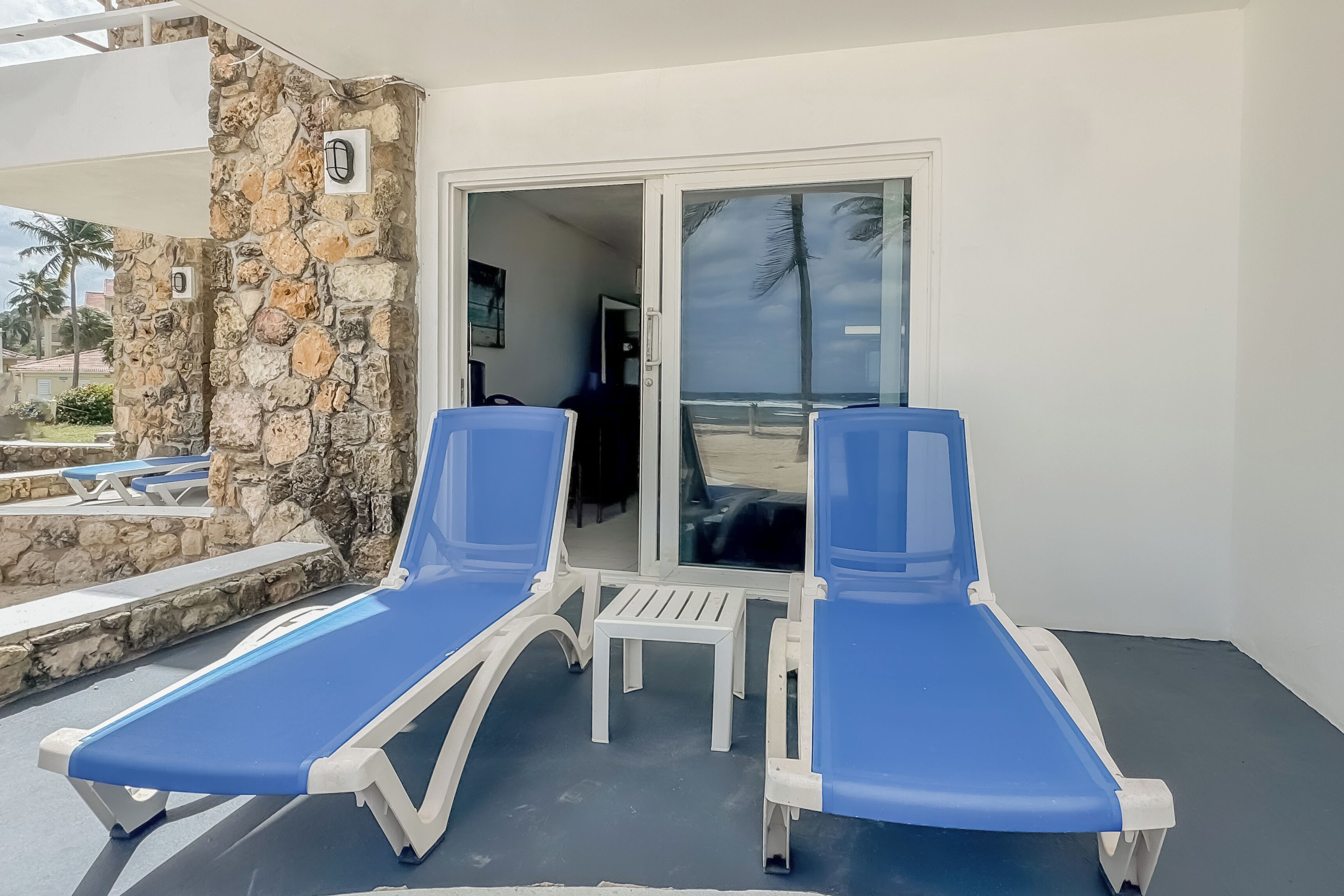 A balcony with two blue lounge chairs and a small white table, overlooking a beach and palm trees.