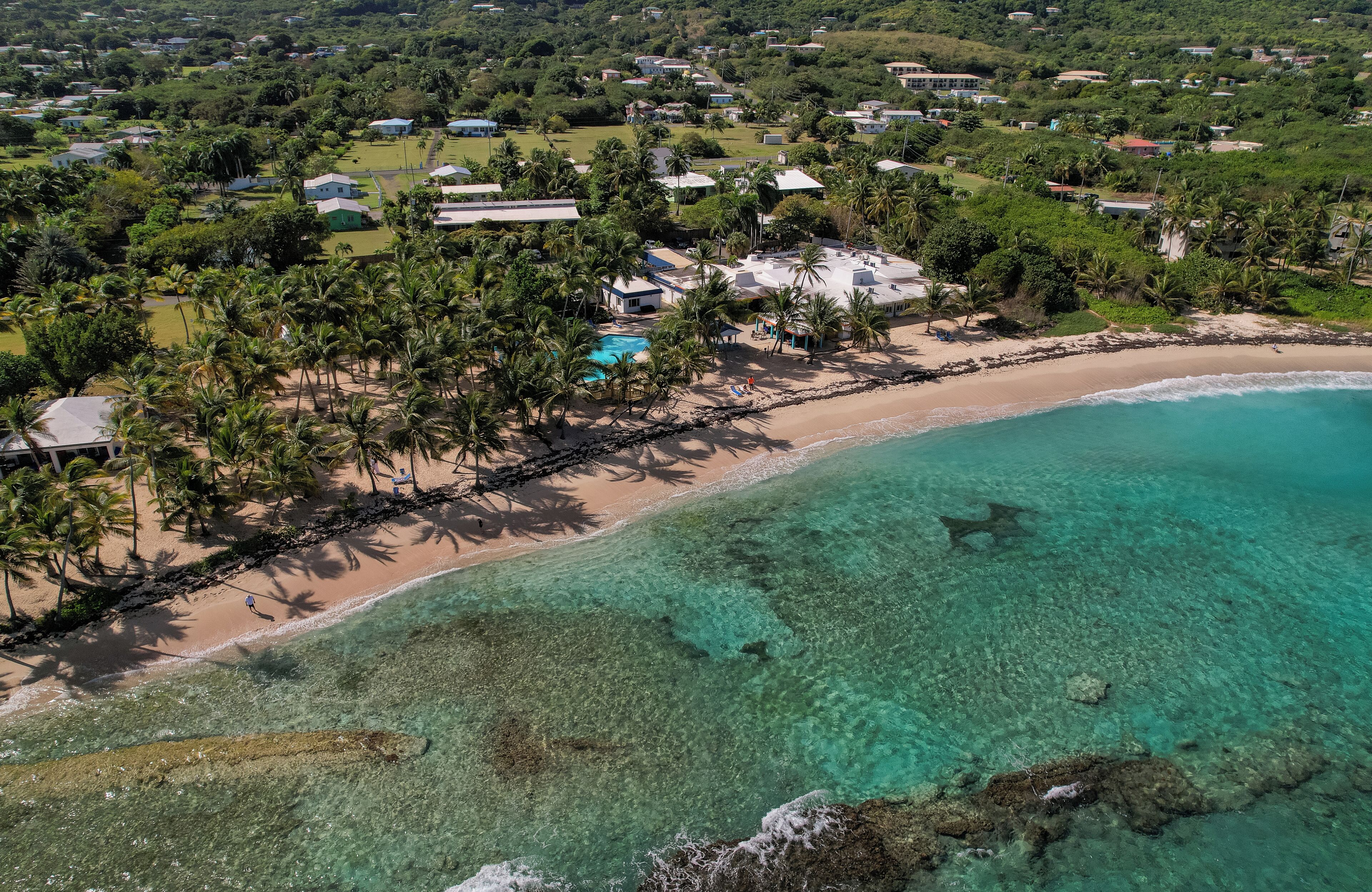 Vista aérea de um resort à beira-mar com águas turquesas cristalinas, uma praia arenosa e diversas palmeiras.