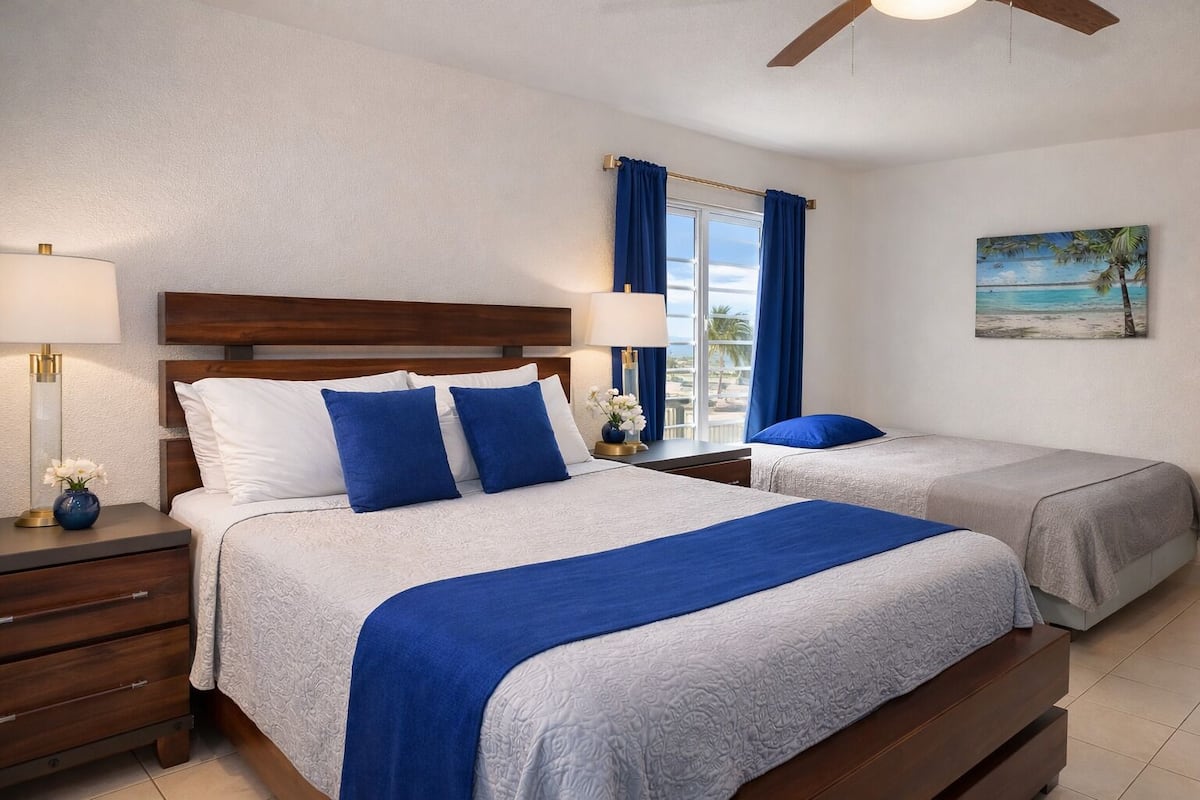 Une chambre d’hôtel avec deux lits, une table de chevet avec une lampe, une fenêtre avec des rideaux et un tableau représentant une scène de plage.