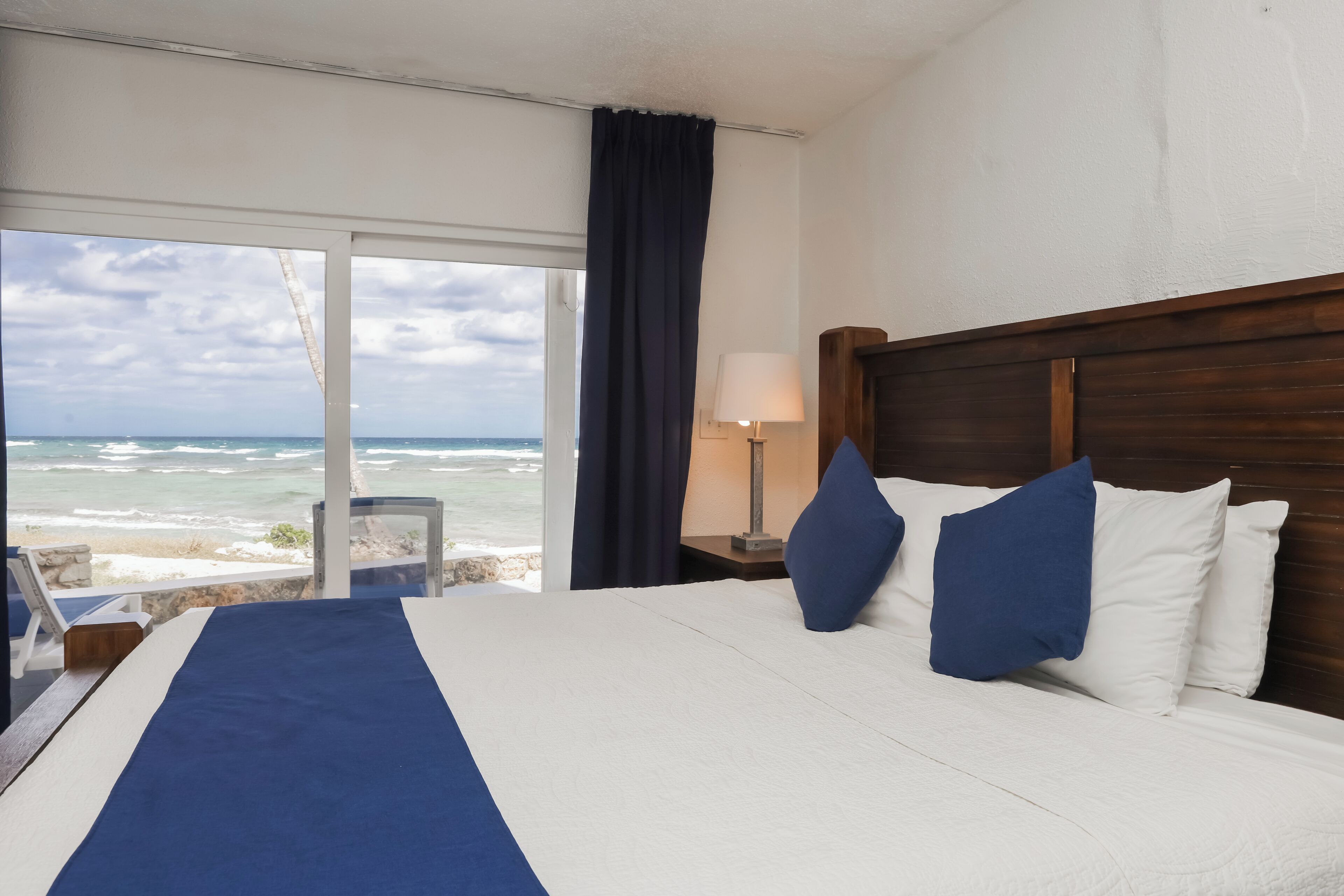 A hotel room with a large bed, a nightstand, a lamp, and a view of the ocean.