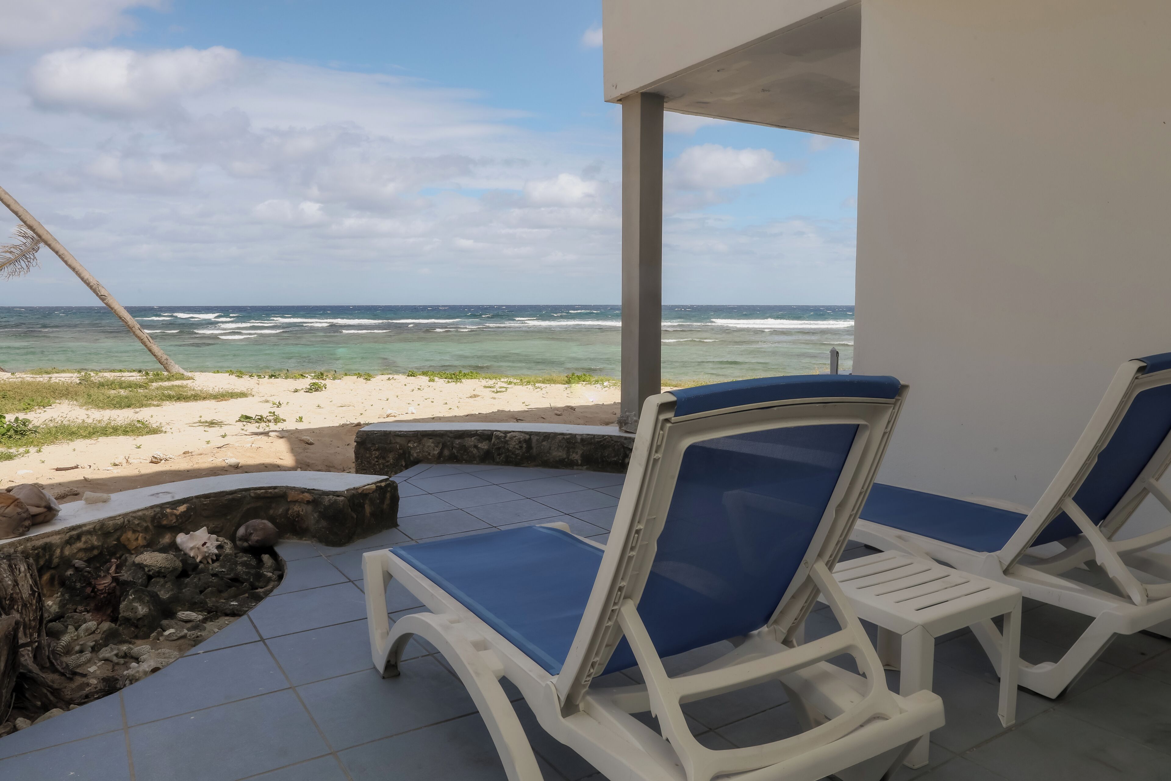 A beachside lounge with two white lounge chairs with blue cushions, a stone wall, and a view of the ocean.