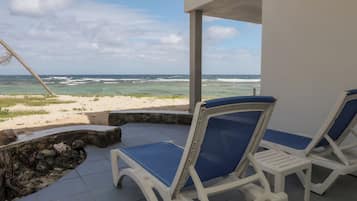 A beachside lounge with two white lounge chairs with blue cushions, a stone wall, and a view of the ocean.