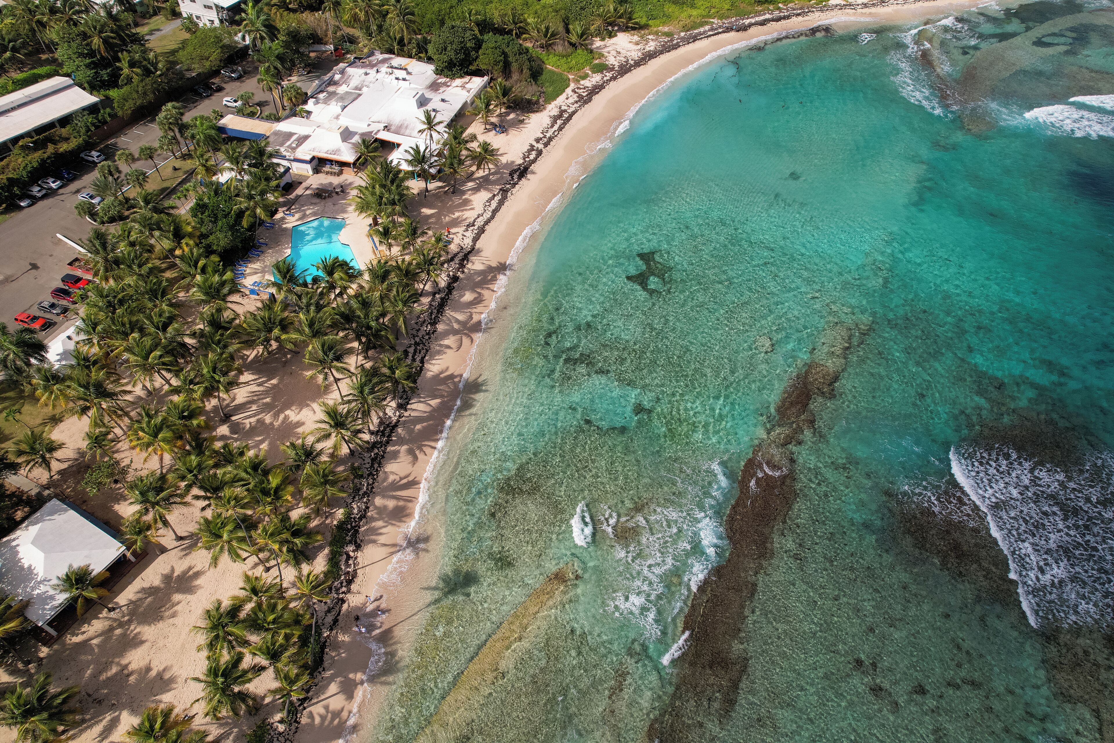 An aerial view of a beach resort with clear turquoise waters, a swimming pool, and palm trees.