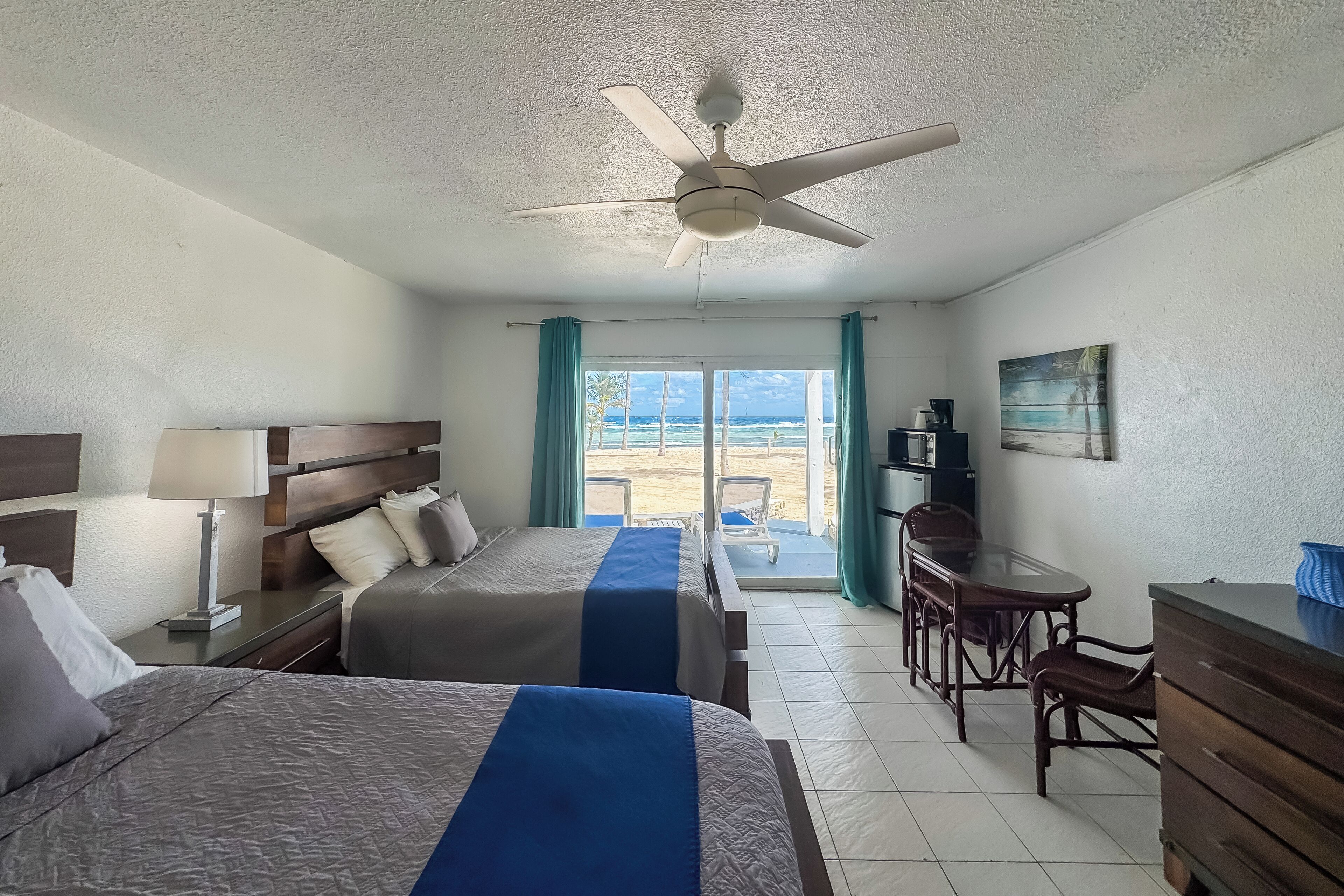 Een hotelkamer met twee bedden, een bureau, een stoel, een plafondventilator, een raam met uitzicht op het strand en een schilderij aan de muur.