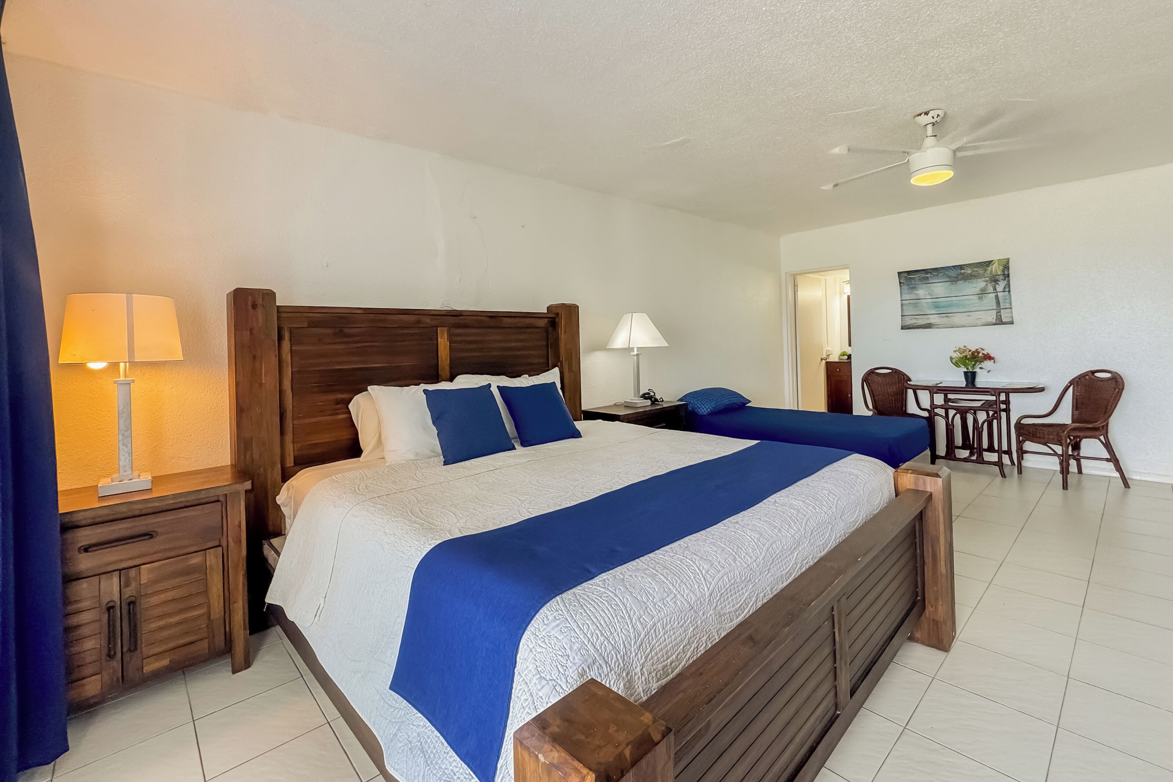 A double bed with a wooden headboard, two bedside tables with lamps, a small table with chairs, and a ceiling fan.