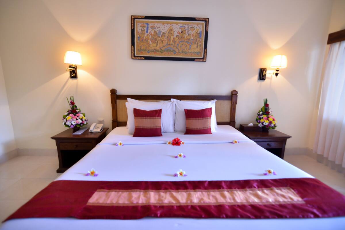 Una habitación de hotel con una cama grande, dos mesitas de noche con lámparas, un arreglo floral en cada una y un cuadro en la pared.