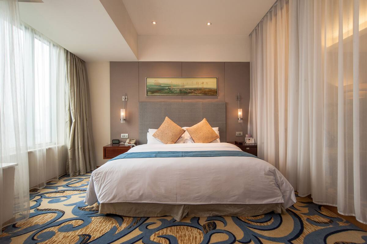 Een hotelkamer met een groot bed, een hoofdbord met een schilderij, twee nachtkastjes met lampen en een kamerbreed tapijt met dessin.