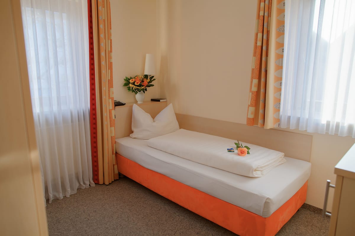 A single bed with white and orange bedding, a bedside table with a lamp, and a vase with flowers.