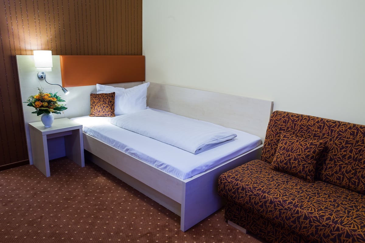 A hotel room with a bed, a nightstand with a lamp, a vase of flowers, and a patterned armchair.